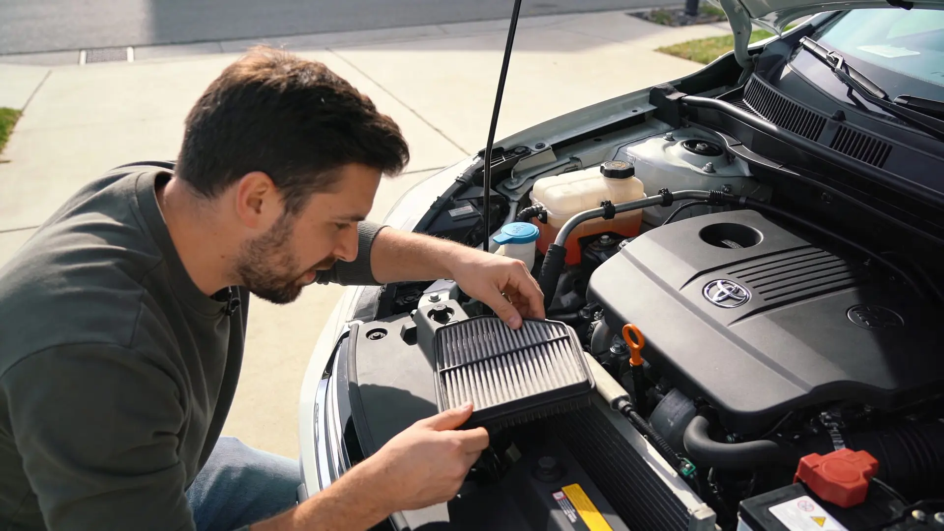 A man inspecting a dirty automotive air filter while performing routine car engine maintenance in a driveway.