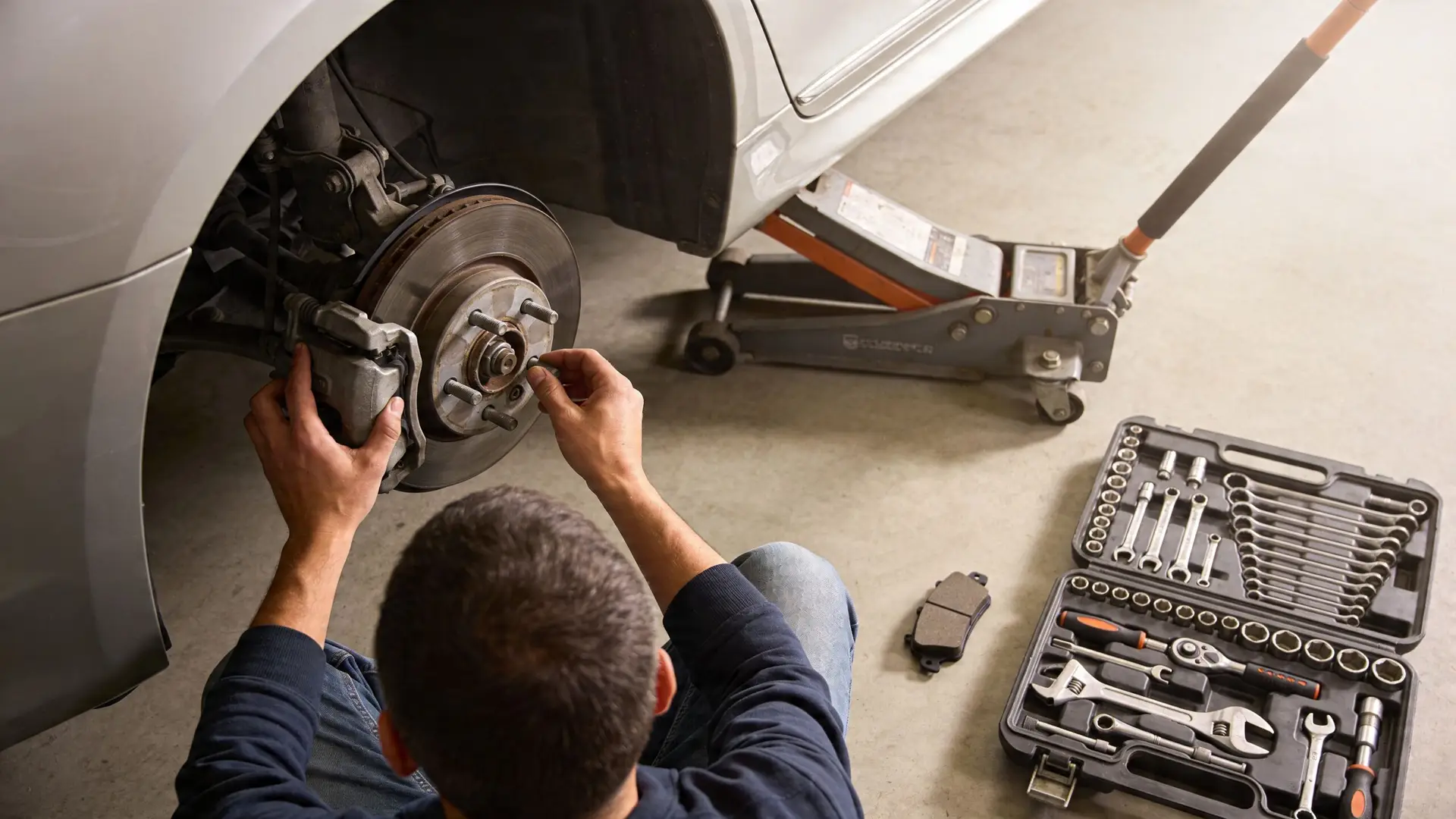 A mechanic working on the brake system of a silver car lifted by a jack in a garage.
